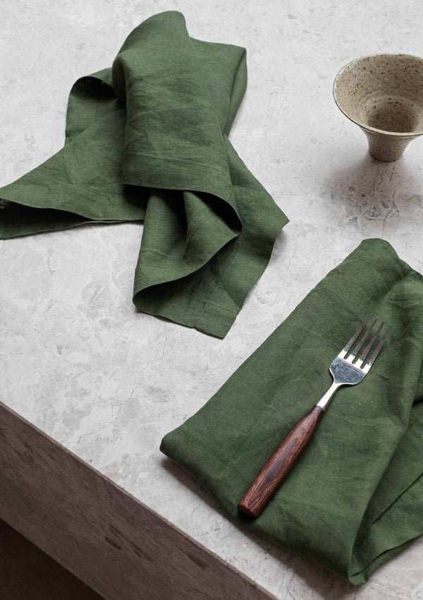 Forest green linen napkins with a wooden fork and ceramic bowl on stone surface