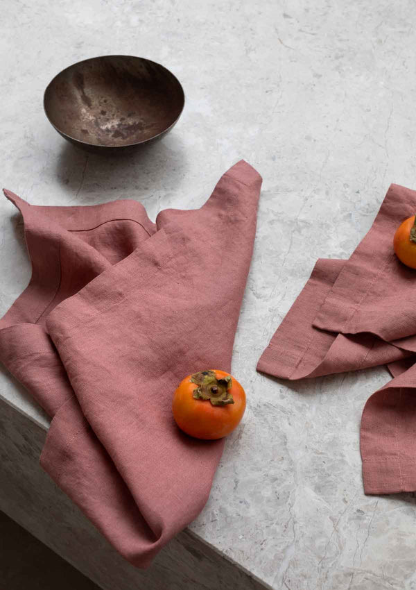 Rose linen napkins on a stone surface, next to a small bowl and persimon fruit.