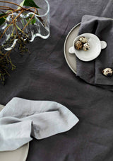 Graphite grey and cool grey linen napkins on a linen tablecloth with pottery dishes, spotted minature eggs and foliage.