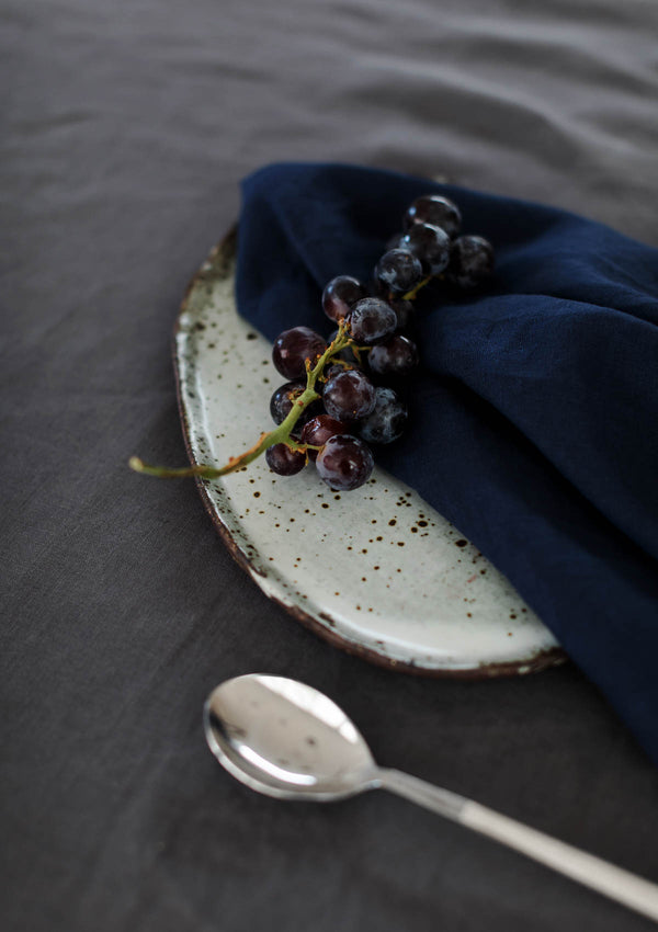 Linen navy napkin on a grey linen tablecloth with a ceramic plate, silver spoon and purple grapes.
