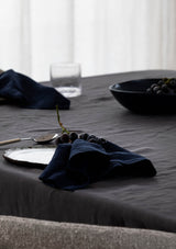 Linen navy napkins on a grey linen tablecloth  alongside a ceramic bowl and plate and purple grapes.