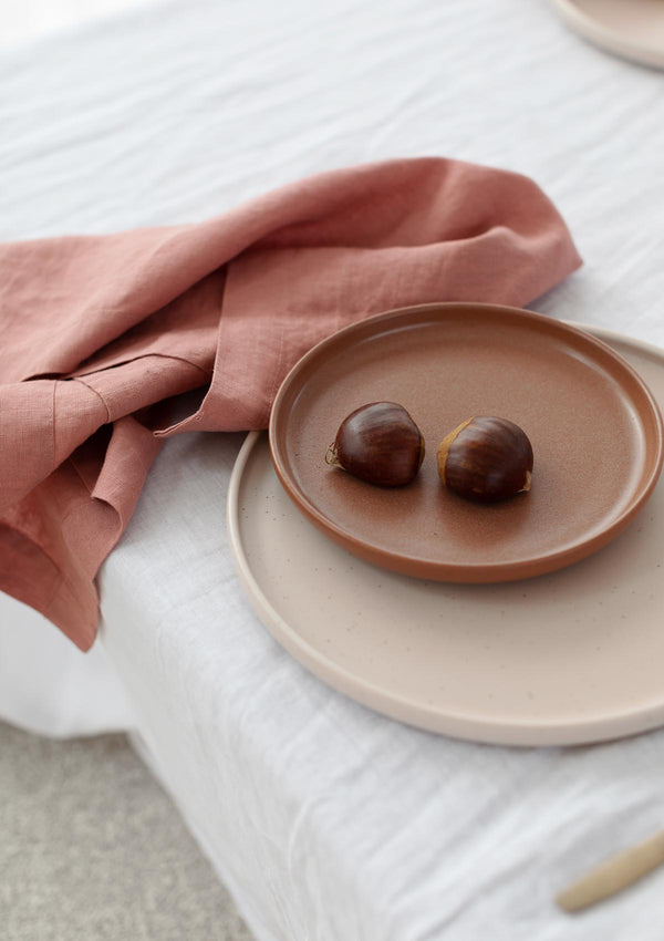 Rose linen napkins on  white tablecloth alongside ceramic plates and chestnuts. 